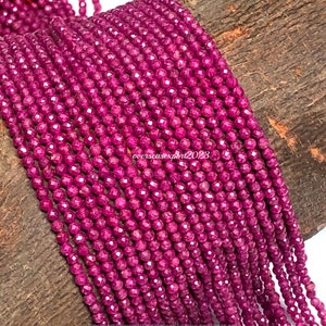 May include: A strand of deep pink ruby beads, each bead is faceted and round, the beads are strung on a clear thread, the beads are displayed on a brown wooden surface.