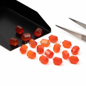 Puede incluir: Gemas facetadas de color rojo anaranjado dispersas sobre una superficie blanca, algunas en una bandeja negra. Se ve un par de pinzas. El texto "Overseasexport2023" está presente. Adecuado para la creación de joyas.