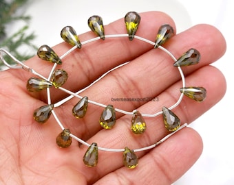 May include: A strand of olive-green, teardrop-shaped beads, each faceted for sparkle, strung on silver wire. The beads, with a mix of light and dark green tones, are displayed against a neutral background. Each bead is approximately 1 cm in length.