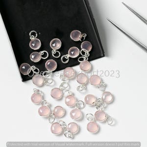 May include: A selection of pink gemstone pendants with silver loops. The polished, round stones have a soft, translucent appearance. The pendants are scattered on a black surface, with silver tools in the background. The text "epert2023" is visible.