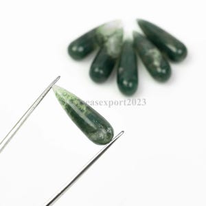 Puede incluir: Una colección de seis piedras preciosas de ágata musgo, lisas y con forma de lágrima. Una piedra verde pulida con inclusiones blancas es sostenida por unas pinzas de metal plateado, mientras que cinco piedras idénticas se disponen en semicírculo.