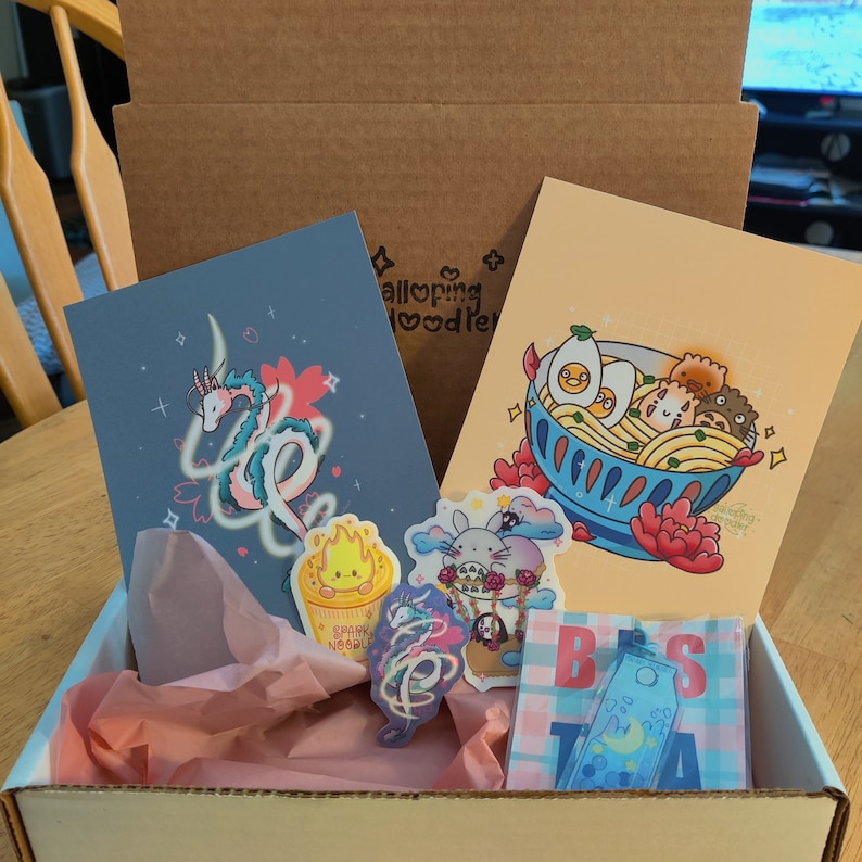 May include: A cardboard box filled with a variety of items, including a blue and white illustration of a dragon, a pink and white illustration of a bunny, a yellow and white illustration of a noodle bowl, and a blue and white illustration of a moon. The box also contains a pink and white tissue paper and a blue and white card with the text "Best Tea".