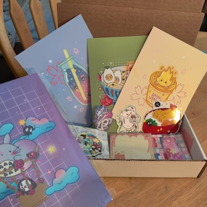 May include: A box filled with various illustrated prints and stickers. The prints feature a glass bottle with stars, a bowl of ramen noodles, and a cup of instant noodles. The stickers feature a girl with a flower crown, a Totoro character, and a bowl of rice with a fried shrimp.