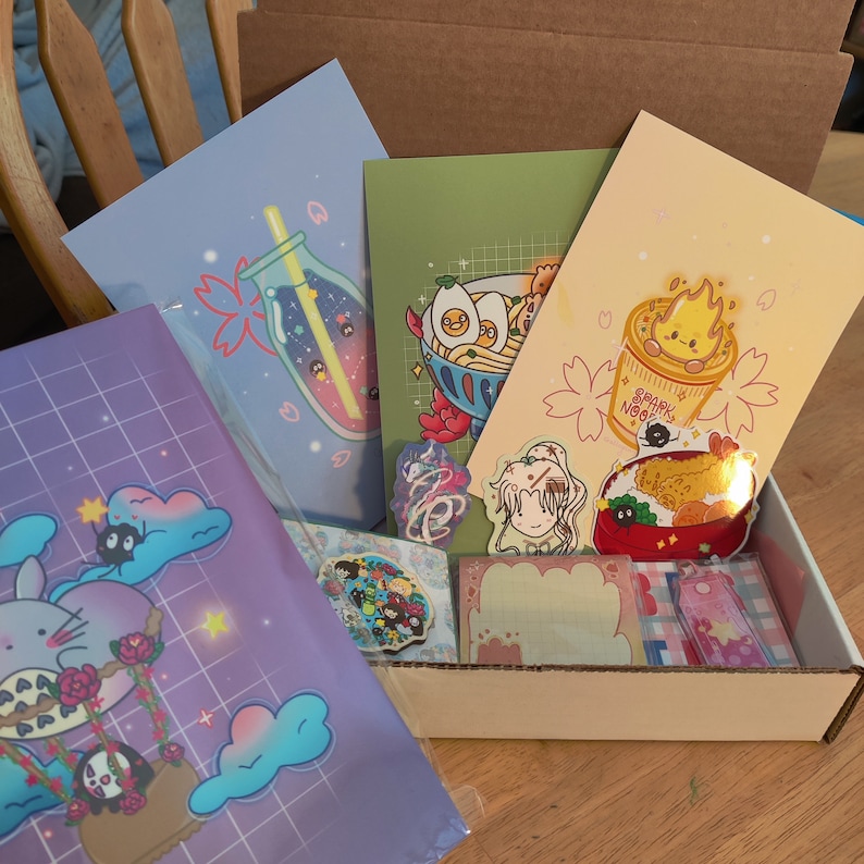 May include: A box filled with various illustrated prints and stickers. The prints feature a glass bottle with stars, a bowl of ramen noodles, and a cup of instant noodles. The stickers feature a girl with a flower crown, a Totoro character, and a bowl of rice with a fried shrimp.