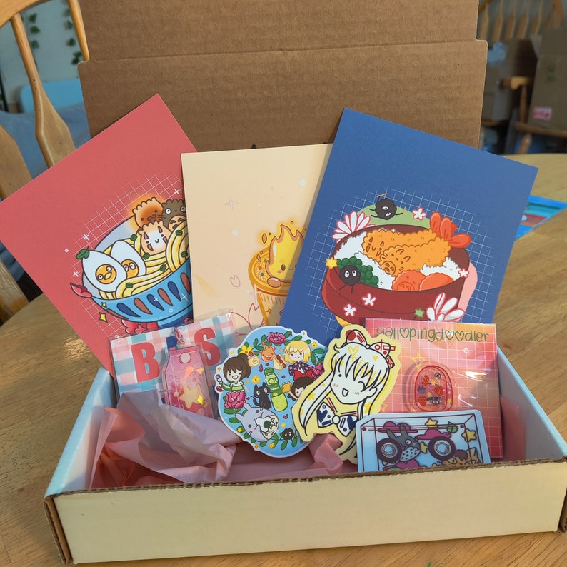 May include: A cardboard box filled with various items, including colorful illustrations, stickers, and pins. The box is open and the items are visible. The illustrations feature anime characters and food. The text "#lopingdoodler" is visible on one of the illustrations.
