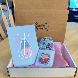 May include: A white cardboard box with a pink tissue paper lining. The box contains a blue and pink illustrated print of a bottle with a straw, a sticker of a group of characters, and a sticker of a bowl of food.