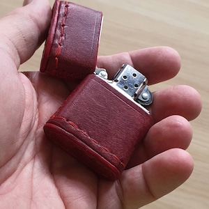 Puede incluir: Una funda de cuero roja para encendedor con un encendedor plateado en el interior. La funda tiene costuras alrededor de los bordes.