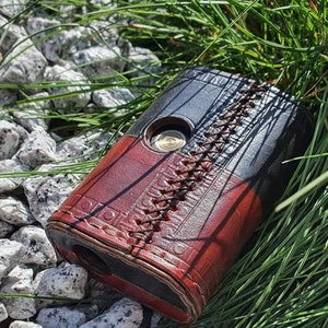 May include: A black and red leather wallet with a stitched design and a circular metal button. The wallet is lying on a bed of small white stones and green grass.