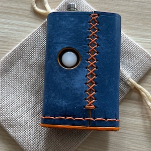 Può includere: Una fiaschetta in pelle blu con cuciture arancioni e un cerchio bianco sul davanti. La fiaschetta è appoggiata su una sacca di tessuto beige.