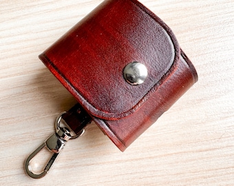 May include: A small, dark red leather case with a silver button and a metal hook. The case is designed to hold small items, such as keys or a phone.