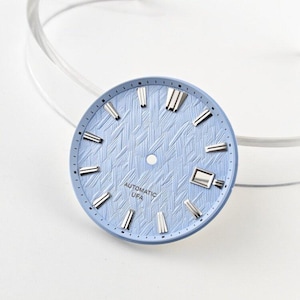 Puede incluir: Una esfera de reloj redonda de color azul claro con un patrón texturizado de líneas entrecruzadas. Incluye índices de bastón aplicados en tono plateado, una ventana de fecha a las tres y el texto "AUTOMATIC" y "UFA" impreso en negro bajo el centro.