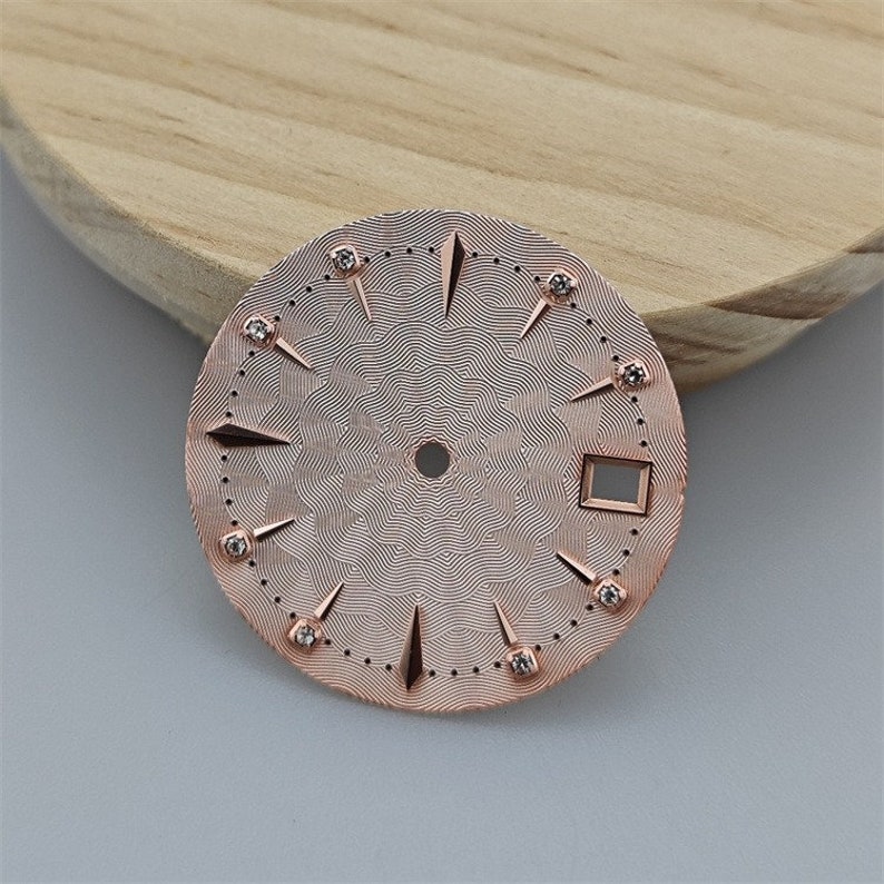 K&ouml;nnte beinhalten: Ein ros&eacute;goldenes Zifferblatt mit einem dekorativen Muster und Diamant-Akzenten. Das Zifferblatt ist f&uuml;r eine Uhr mit einem quadratischen Datumsfenster konzipiert.