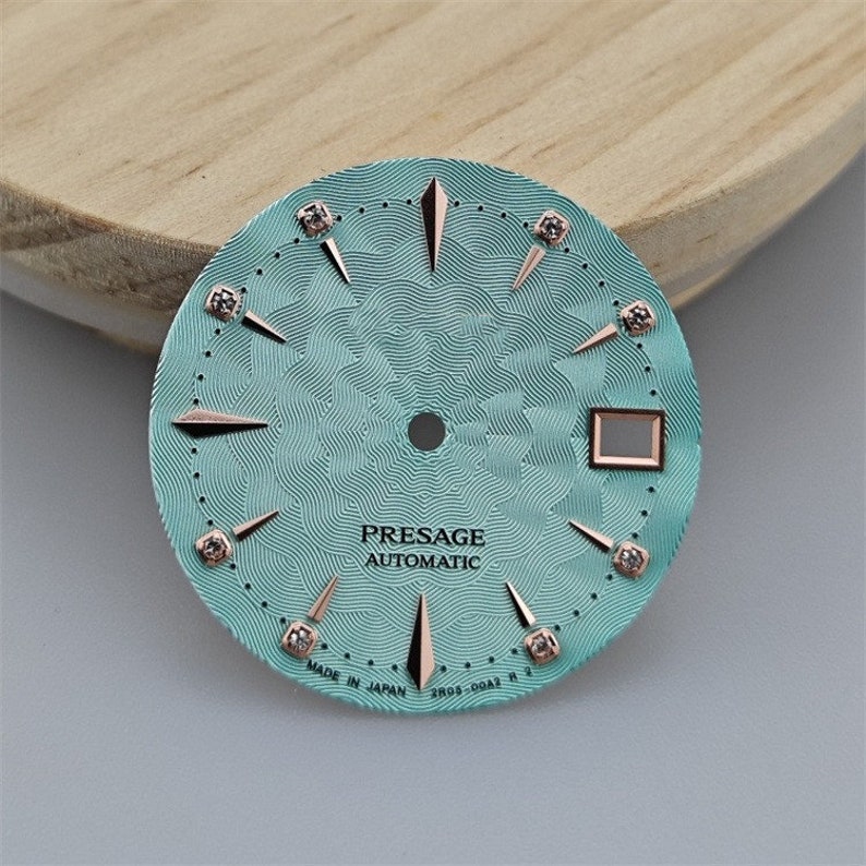 K&ouml;nnte beinhalten: Ein t&uuml;rkisfarbenes Zifferblatt mit ros&eacute;goldenen Akzenten und Diamant-Stundenmarkierungen. Das Zifferblatt zeigt "PRESAGE AUTOMATIC" und "MADE IN JAPAN SR05-00A2 R" an.