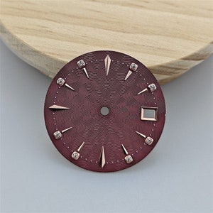 K&ouml;nnte beinhalten: Ein burgunderfarbenes Zifferblatt mit ros&eacute;goldenen Akzenten und Diamant-Stundenmarkierungen. Das Zifferblatt hat ein strukturiertes Muster und ein quadratisches Fenster f&uuml;r das Datum.