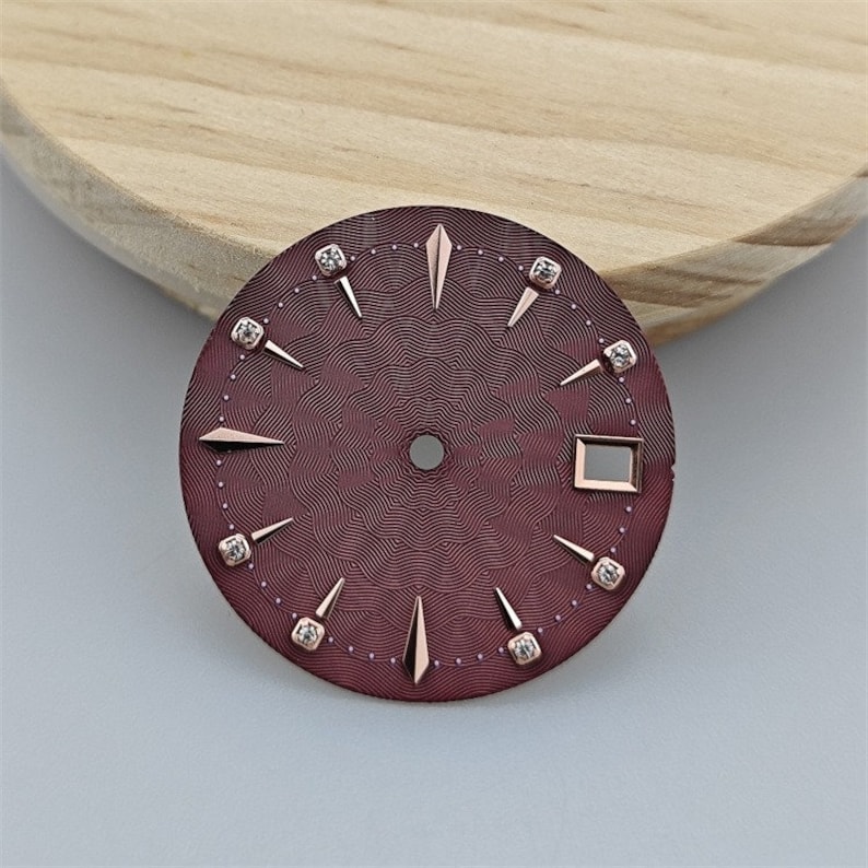 K&ouml;nnte beinhalten: Ein burgunderfarbenes Zifferblatt mit ros&eacute;goldenen Akzenten und Diamant-Stundenmarkierungen. Das Zifferblatt hat ein strukturiertes Muster und ein quadratisches Fenster f&uuml;r das Datum.