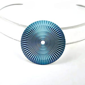Puede incluir: Un colgante circular azul con un patrón radial facetado similar a un sol y un pequeño orificio central, unido a un collar rígido de metal plateado. El colgante presenta tonos azul oscuro y azul claro con líneas geométricas definidas.