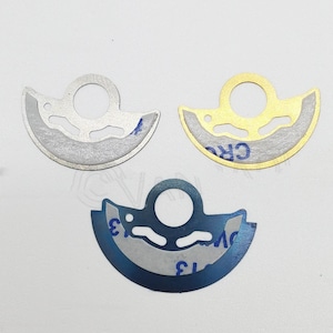 May include: Three metal engine parts in different colors: silver, gold, and blue. Each part has a circular hole in the center and a curved shape.