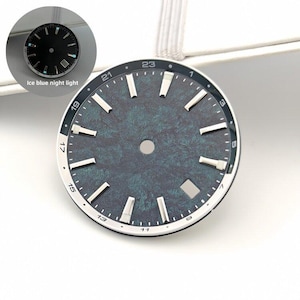 Puede incluir: Una esfera de reloj redonda con una superficie texturizada en verde azulado oscuro, índices de bastón plateados y una ventana de fecha. El borde exterior muestra números impresos del 1 al 23. Un círculo insertado muestra la esfera con luz nocturna azul hielo.