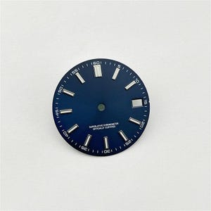 Puede incluir: Una esfera de reloj circular de color azul oscuro con marcadores de hora rectangulares en tono plateado, una ventana de fecha en la posición de las tres y texto impreso en blanco que dice SUPERLATIVE CHRONOMETER OFFICIALLY CERTIFIED.