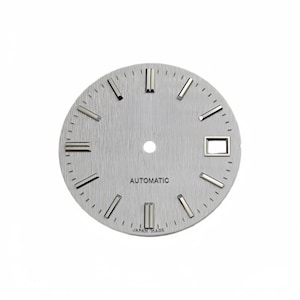 Puede incluir: Una esfera de reloj circular plateada con textura de metal cepillado, que presenta índices de bastón plateados aplicados, una ventana de fecha en la posición de las tres y el texto AUTOMATIC y JAPAN MADE impreso en negro.