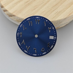 Puede incluir: Una esfera de reloj circular azul con números arábigos orientales plateados en relieve como marcadores de hora. La esfera incluye un orificio central para las manecillas, una ventana de fecha rectangular a las tres y marcas de minutos blancas.