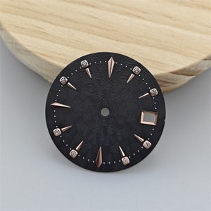 K&ouml;nnte beinhalten: Schwarzes Zifferblatt mit ros&eacute;goldenen Akzenten und Diamant-Stundenmarkierungen. Das Zifferblatt hat ein strukturiertes Muster und ein quadratisches Fenster bei 4 Uhr.