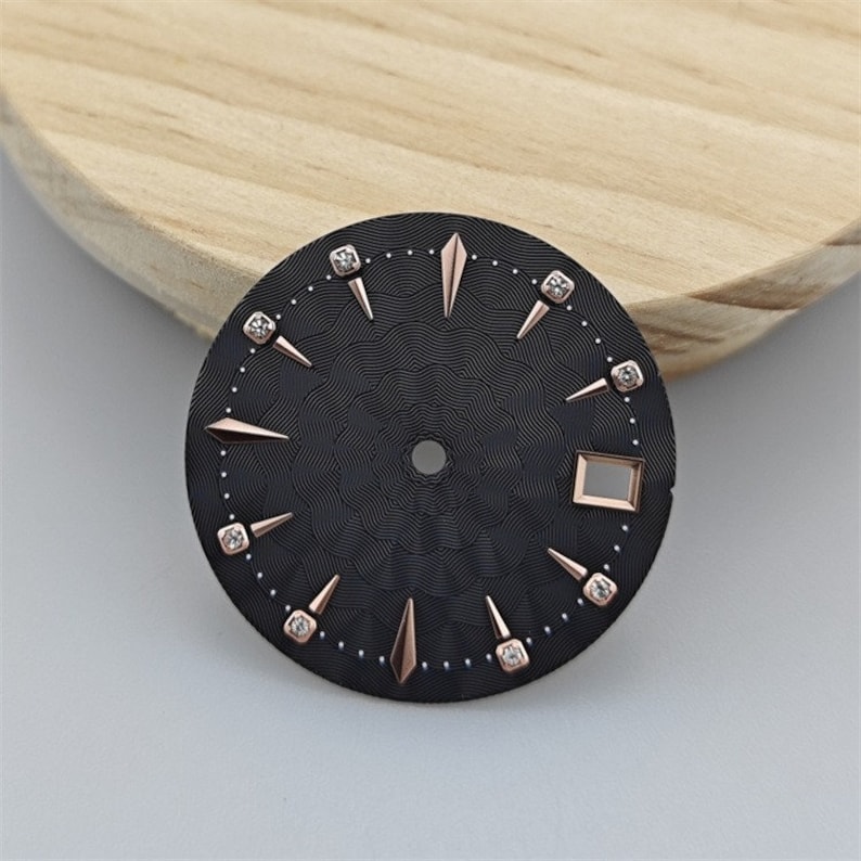 K&ouml;nnte beinhalten: Schwarzes Zifferblatt mit ros&eacute;goldenen Akzenten und Diamant-Stundenmarkierungen. Das Zifferblatt hat ein strukturiertes Muster und ein quadratisches Fenster bei 4 Uhr.