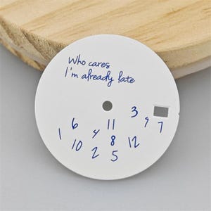 Puede incluir: Una esfera de reloj circular blanca con el texto manuscrito en azul Who cares I'm already late en la parte superior. La esfera muestra números manuscritos en azul dispersos del 1 al 12 en orden no secuencial e incluye una ventana de fecha.