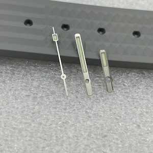 May include: Three watch hands, one long, one medium, and one short, are laid out on a grey surface. The hands are silver with a luminous green fill.