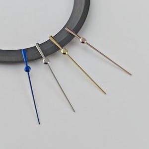 Puede incluir: Cuatro manecillas de reloj delgadas dispuestas en fila, cada una con un punto central circular. Las manecillas tienen acabados metálicos distintos: azul profundo, plata pulida, oro brillante y un tono oro rosa cálido, cerca de un anillo oscuro.