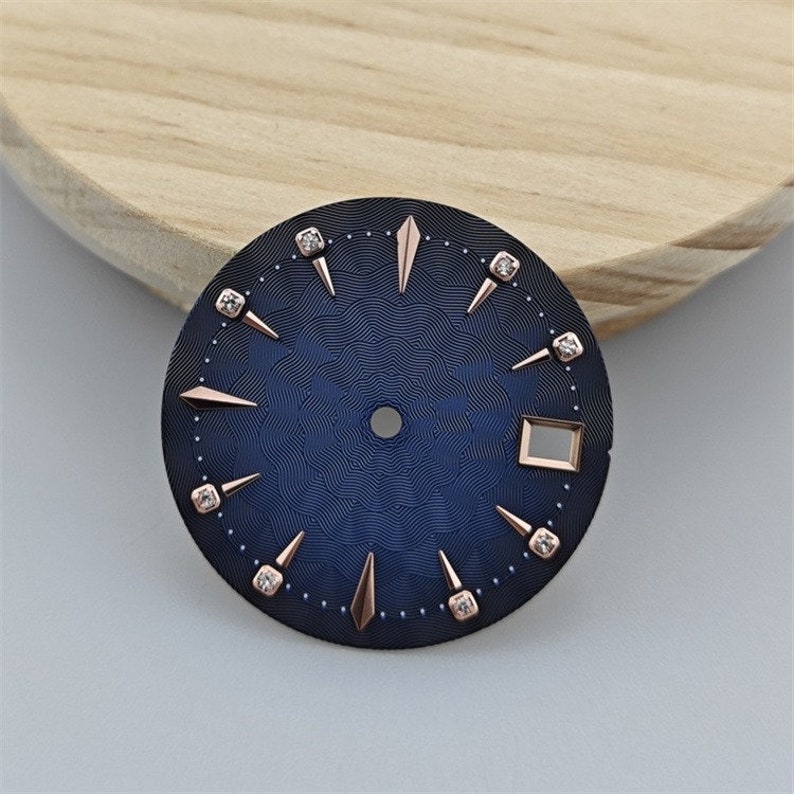 K&ouml;nnte beinhalten: Ein blaues Zifferblatt mit ros&eacute;goldenen Akzenten und Diamant-Stundenmarkierungen. Das Zifferblatt hat ein strukturiertes Muster und ein quadratisches Datumsfenster.