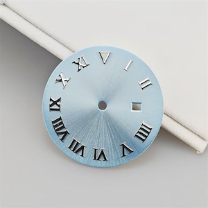 May include: A circular watch dial with a light blue sunburst finish, featuring applied silver-tone Roman numeral hour markers. The dial has a central hole for hands and a rectangular date window aperture positioned at the three o'clock marker.