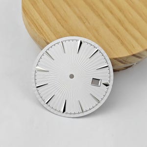 Peut inclure: Un cadran de montre circulaire blanc avec un motif guilloché en rayons de soleil, des index triangulaires argentés et un guichet de date rectangulaire à trois heures. Le cadran présente un trou central pour les aiguilles et une minuterie.
