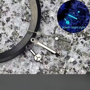 Puede incluir: Un juego de tres manecillas de reloj plateadas que incluye una manecilla larga y delgada, una manecilla cónica más ancha y una manecilla pequeña con base circular. Un círculo muestra las manecillas brillando con luz azul BGW9- Night Light.