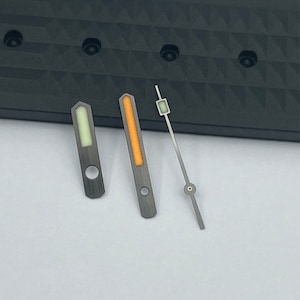 May include: Three watch hands, one with a green luminescent stripe, one with an orange stripe, and one with a silver rectangle. The hands are on a black background.