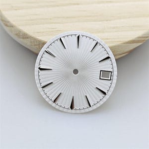 Puede incluir: Una esfera de reloj circular plateada con un patrón de rayos de sol y líneas radiantes. Incluye marcadores de hora negros aplicados, un orificio central para las manecillas y una ventana de fecha rectangular en la posición de las tres.