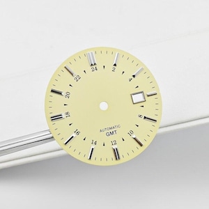 Op de afbeelding: Een ronde, lichtgele wijzerplaat met zilverkleurige rechthoekige uurmarkeringen, een centrale ronde opening en zwarte cijfers voor een 24-uurs schaal. De tekst "AUTOMATIC" en "GMT" is in het zwart boven het middelpunt gedrukt.