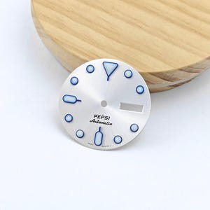 Puede incluir: Una esfera de reloj circular plateada con acabado de rayos de sol, con marcadores de hora de borde azul y centros luminosos blancos. La esfera muestra el texto PEPSI Automatic en negro, un orificio central para las manecillas y una ventana de fecha.