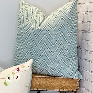May include: A blue and white chevron patterned throw pillow with a subtle sheen. The pillow is sitting on a woven wicker surface.