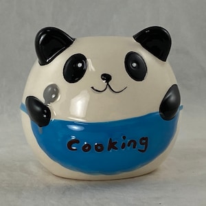 May include: A white ceramic panda figurine with black ears, eyes, and nose. The panda is wearing a blue apron with the word "Cooking" written on it.