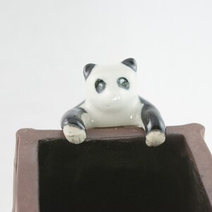 May include: A brown ceramic planter with a black panda bear figurine peeking over the edge. The panda has black eyes and a white face.