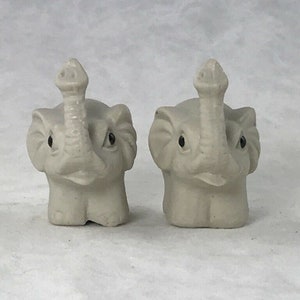 May include: Two small, white ceramic elephant figurines with black eyes and raised trunks. The elephants are facing forward and have a simple, stylized design.