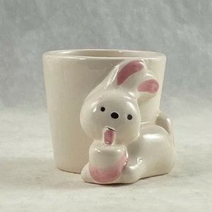 May include: A white ceramic planter shaped like a bunny with pink ears and nose. The planter is sitting on a white surface.