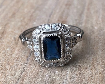 May include: A silver ring with a dark blue square-shaped gemstone surrounded by smaller clear gemstones.