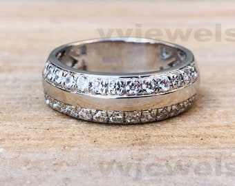 Puede incluir: Un anillo plateado con dos filas de pequeñas piedras preciosas transparentes y una banda central más ancha. El anillo se muestra sobre una superficie de madera marrón claro, resaltando su diseño y brillo.