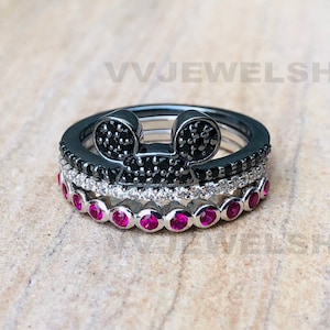May include: Three stacked rings. The top ring is black with a Mickey Mouse head design made of black stones. The middle ring is silver with white stones. The bottom ring is silver with red stones. The rings are on a light brown surface.