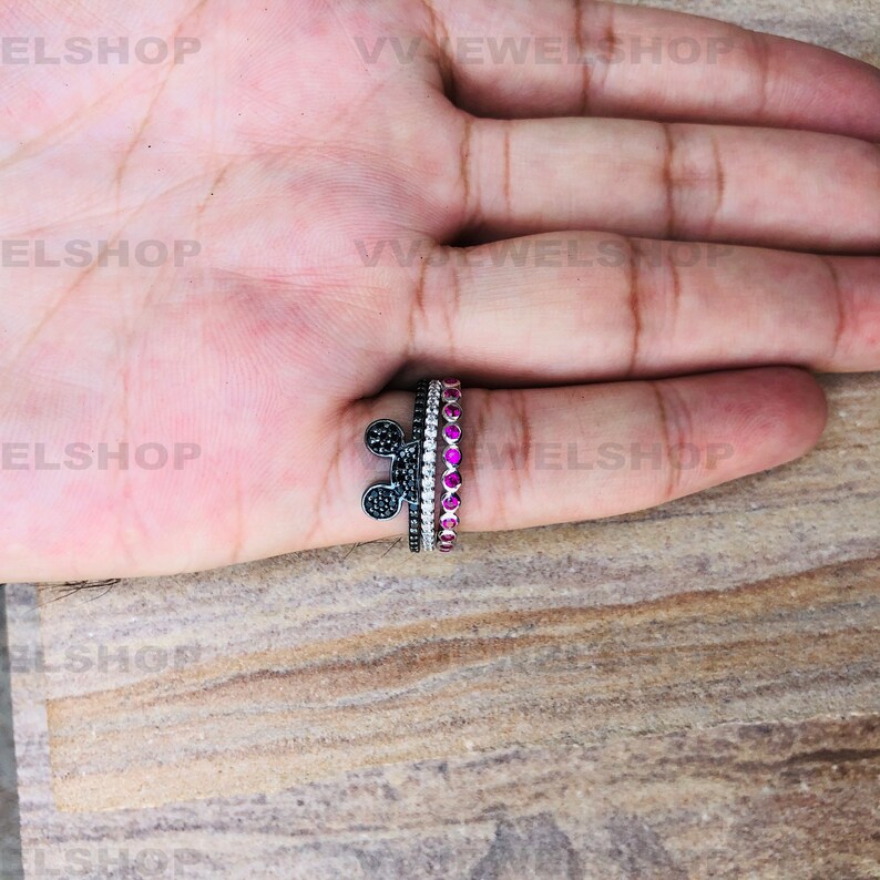 May include: Three rings on a finger. One ring features a black Mickey Mouse head design, another has white stones, and the third has pink stones. The rings are set against a light brown wooden surface.