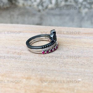 May include: A stack of three rings. The top ring is black with a decorative design. The middle ring is silver with black stones. The bottom ring is silver with red stones. The rings are on a light brown surface.
