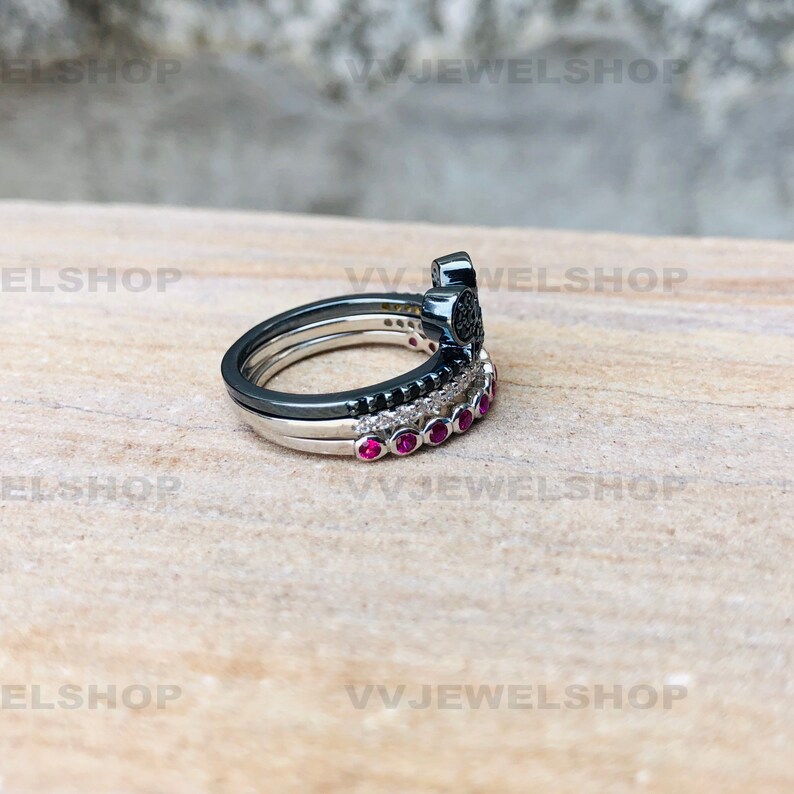 May include: A stack of three rings. The top ring is black with a decorative design. The middle ring is silver with black stones. The bottom ring is silver with red stones. The rings are on a light brown surface.