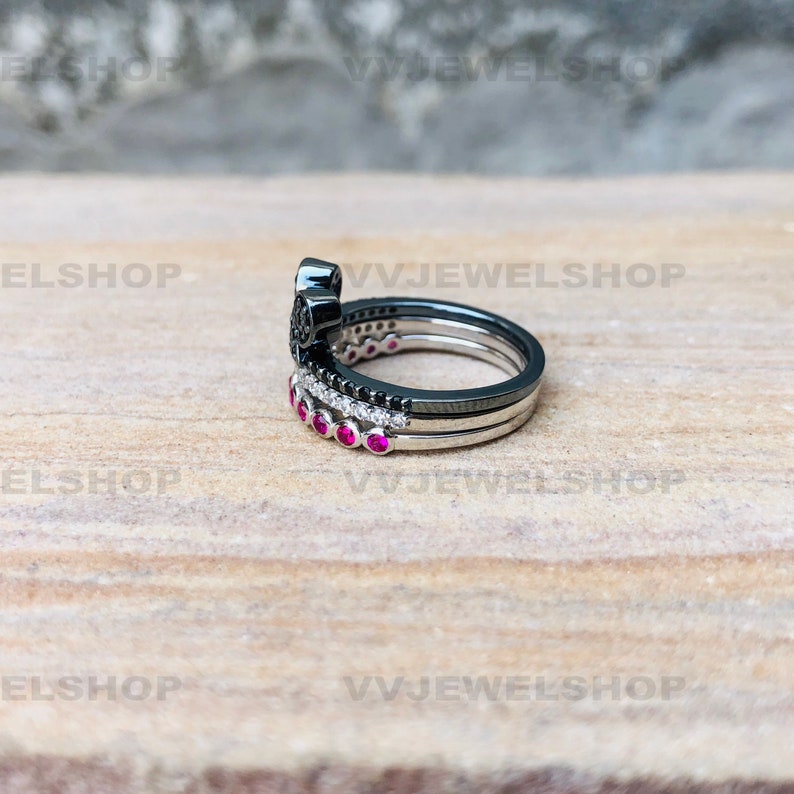 May include: A stack of three rings with a black, silver, and white color scheme. The top ring has a black floral design. The middle ring has black and white stones. The bottom ring has pink stones. The rings are on a light brown surface.